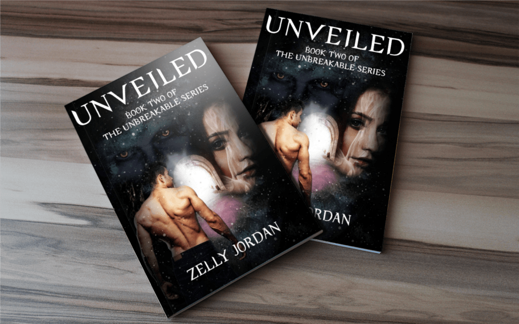 Two copies of the book 'Unveiled' by Zelly Jordan, displayed on a wooden surface. The cover features a male figure and a female figure surrounded by a cosmic background.