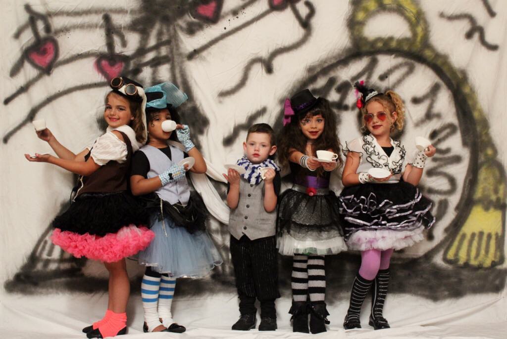 A group of five children dressed in colorful costumes, featuring frilly skirts and unique accessories, posing playfully with small teacups against a whimsical backdrop.