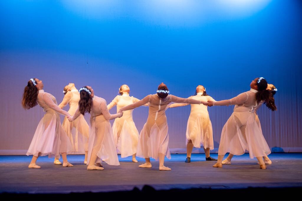 A group of dancers in white dresses performing on stage, holding hands in a circle, with a blue backdrop and soft lighting.